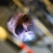 A close-up, low-angle photograph of a man's face reflected or viewed through a glass or transparent surface, illuminated by a bright light source. The image is taken from an unusual perspective, likely looking up through a glass container or optical element. The composition creates a distorted, artistic view of the subject examining or inspecting something closely.