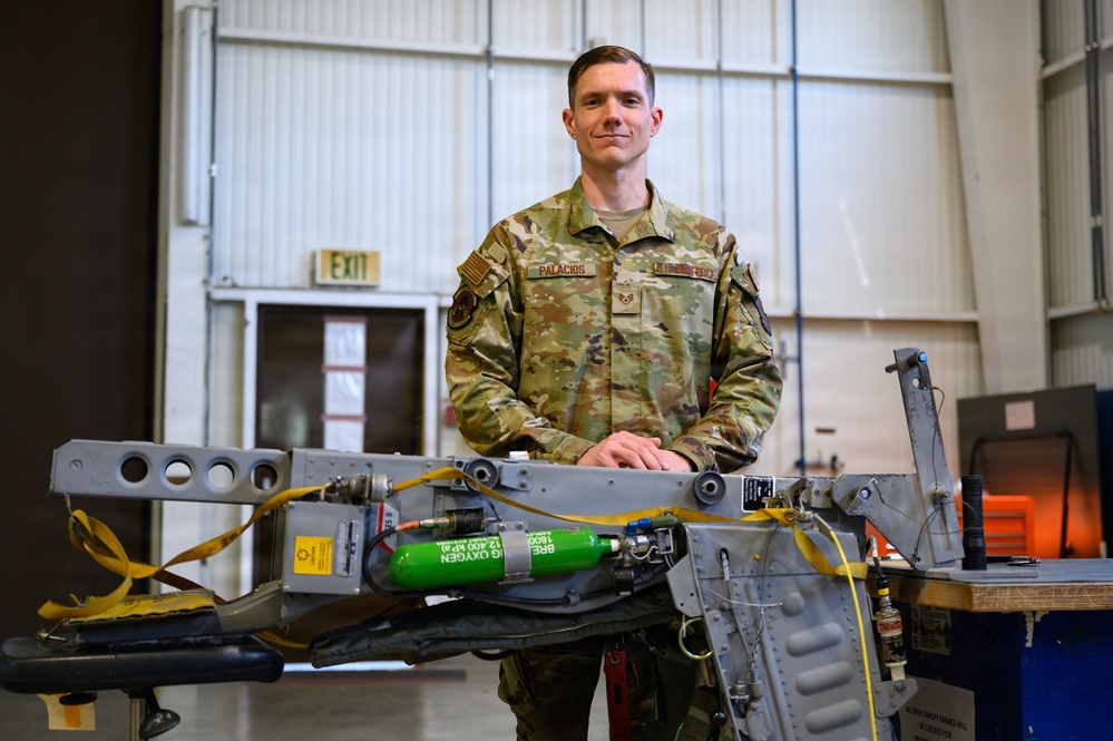 A U.S. Air Force service member named Palacios, wearing OCP (Operational Camouflage Pattern) uniform, poses in what appears to be an aircraft maintenance hangar. He is standing behind a piece of aircraft equipment that includes green oxygen cylinders and yellow safety straps. The setting suggests a professional military maintenance environment.