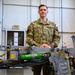 A U.S. Air Force service member named Palacios, wearing OCP (Operational Camouflage Pattern) uniform, poses in what appears to be an aircraft maintenance hangar. He is standing behind a piece of aircraft equipment that includes green oxygen cylinders and yellow safety straps. The setting suggests a professional military maintenance environment.