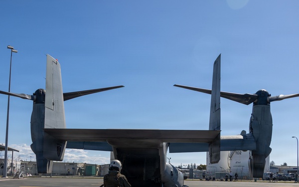 A military tiltrotor aircraft, identified as a Bell Boeing V-22 Osprey (tail number 702), is parked on a tarmac at what appears to be a military airfield or base. A single crew member in flight gear and helmet stands at the rear ramp of the aircraft, facing the open cargo bay. The setting appears to be a sunny day at a well-maintained facility with light poles, buildings, and other personnel visible in the background.