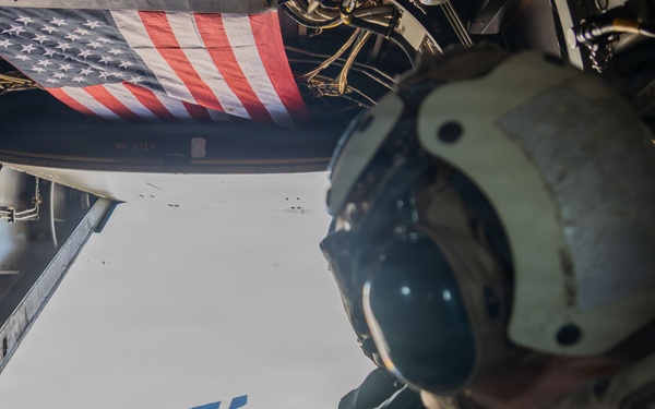 A military service member wearing a flight helmet and camouflage uniform is seated at the open rear ramp of a military aircraft in flight. An American flag is visible attached to the interior of the aircraft above the opening. The perspective suggests the photo was taken from inside the aircraft looking outward toward another aircraft visible in the distance.