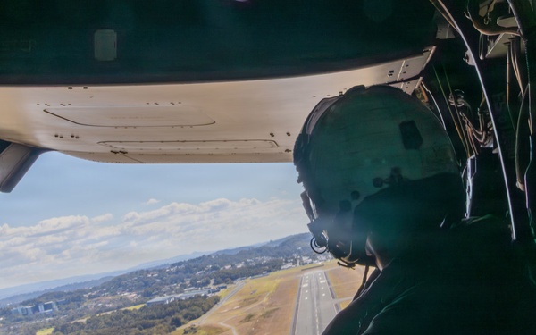 A crew member wearing a flight helmet and headset is seated inside a military aircraft, looking out through a large rear cargo ramp opening toward an airport runway below. The aircraft appears to be on approach or low-altitude flight over a suburban/wooded area. Weight limit placards are partially visible on the interior wall to the right.