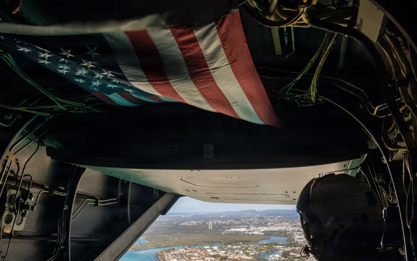 A view from inside a military helicopter, likely a CH-47 Chinook, showing a crew member in flight gear looking out through the open rear ramp at a coastal city below. An American flag is affixed to the interior ceiling of the aircraft. The aerial perspective reveals a waterfront urban area with buildings, a beach, and a river or inlet.