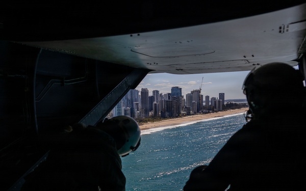 Two military personnel in flight helmets are seated at the open rear ramp of a military aircraft, looking out over a coastal city with a sandy beach and turquoise water below. The image is taken from inside the aircraft looking outward, creating a strong silhouette effect against the bright exterior. A skyline of high-rise buildings is visible in the background along the coastline.