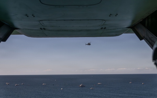 The image is taken from inside a large military aircraft, looking out through an open rear cargo ramp or door toward the ocean below. Two helmeted crew members are visible in silhouette on either side of the frame, observing a scene with numerous boats clustered on the water's surface. A smaller helicopter is visible in the mid-distance sky above the boats.