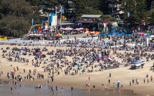 An aerial view of a crowded beach event with hundreds of attendees gathered on the sand. A large organized event is taking place with branded tents, stages, and vendor areas visible along the beachfront. The Red Bull branding is prominently visible, suggesting a sponsored sporting or entertainment event.