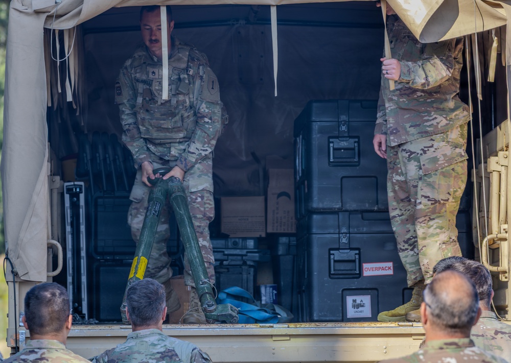 A U.S. Army soldier in OCP (Operational Camouflage Pattern) uniform stands in the back of a military cargo vehicle, handling what appears to be a mortar tube or similar crew-served weapon system. Several other soldiers in the foreground observe what appears to be a demonstration or training event. Military equipment cases, including one labeled 'OVERPACK', are visible in the vehicle's cargo area.