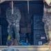 A U.S. Army soldier in OCP (Operational Camouflage Pattern) uniform stands in the back of a military cargo vehicle, handling what appears to be a mortar tube or similar crew-served weapon system. Several other soldiers in the foreground observe what appears to be a demonstration or training event. Military equipment cases, including one labeled 'OVERPACK', are visible in the vehicle's cargo area.