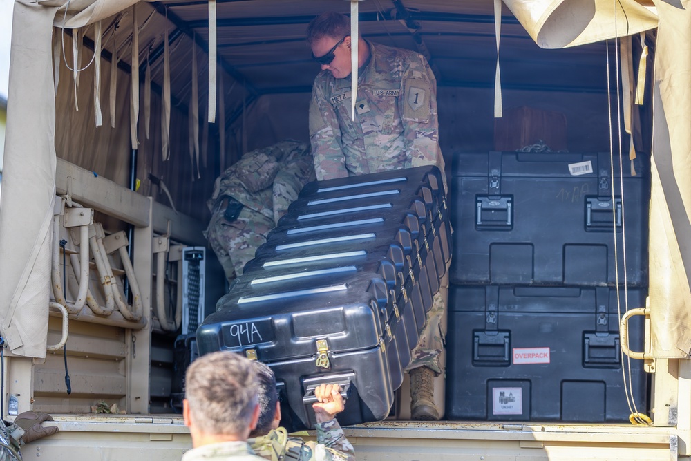 U.S. Army soldiers are loading or unloading equipment cases into the cargo area of a military vehicle. At least two soldiers in OCP (Operational Camouflage Pattern) uniforms are visible, one inside the vehicle bed and one below passing up a hard-shell case marked '94A'. Multiple black hard-shell equipment cases and military storage containers are visible in the cargo area.