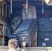 U.S. Army soldiers are loading or unloading equipment cases into the cargo area of a military vehicle. At least two soldiers in OCP (Operational Camouflage Pattern) uniforms are visible, one inside the vehicle bed and one below passing up a hard-shell case marked '94A'. Multiple black hard-shell equipment cases and military storage containers are visible in the cargo area.