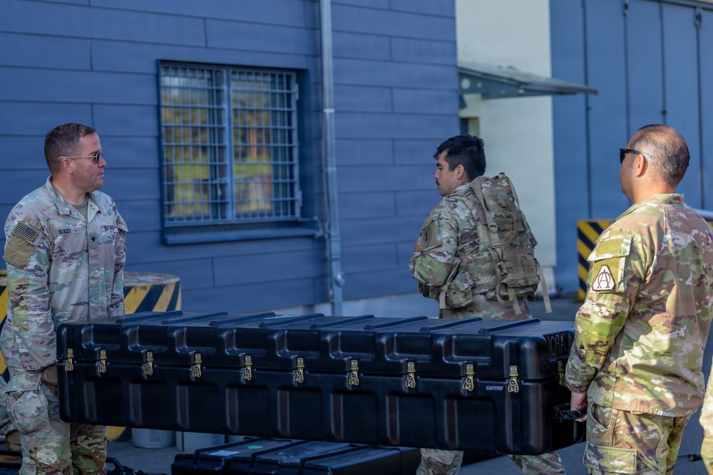 Three U.S. Army soldiers in MultiCam/OCP camouflage uniforms are handling a large black hard-shell equipment case outside a building. Two soldiers are carrying the case while a third observes. The setting appears to be a military installation or facility, identifiable by the building's architecture and safety markings.
