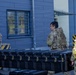 Three U.S. Army soldiers in MultiCam/OCP camouflage uniforms are handling a large black hard-shell equipment case outside a building. Two soldiers are carrying the case while a third observes. The setting appears to be a military installation or facility, identifiable by the building's architecture and safety markings.