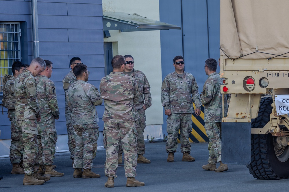 A group of approximately 10 military personnel in US Army OCP (Operational Camouflage Pattern) uniforms are gathered in a briefing or discussion formation near a military vehicle. They are standing on a paved surface adjacent to a building, with a tan-colored military truck (likely an LMTV or FMTV) visible on the right side. The setting appears to be a military installation or staging area.