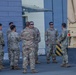 A group of approximately 10 military personnel in US Army OCP (Operational Camouflage Pattern) uniforms are gathered in a briefing or discussion formation near a military vehicle. They are standing on a paved surface adjacent to a building, with a tan-colored military truck (likely an LMTV or FMTV) visible on the right side. The setting appears to be a military installation or staging area.