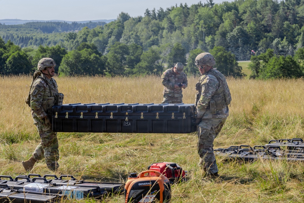Three military personnel in camouflage uniforms and helmets are handling equipment in an open field. Two soldiers are carrying a large black hard-shell case marked '113A', while a third soldier stands nearby. Multiple equipment cases and a Generac generator are visible on the ground around them.
