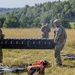 Three military personnel in camouflage uniforms and helmets are handling equipment in an open field. Two soldiers are carrying a large black hard-shell case marked '113A', while a third soldier stands nearby. Multiple equipment cases and a Generac generator are visible on the ground around them.