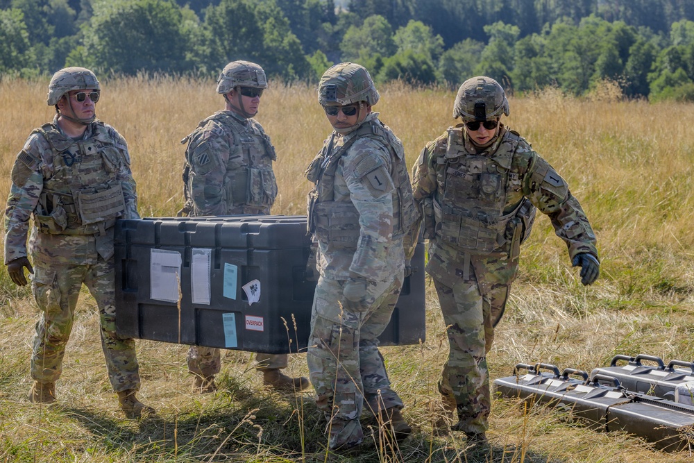 Four military personnel in U.S. Army OCP (Operational Camouflage Pattern) uniforms are carrying a large black equipment case across an open grassy field. Two soldiers are actively transporting the case while two others walk alongside. Additional equipment cases are visible on the ground nearby.