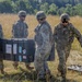 Four military personnel in U.S. Army OCP (Operational Camouflage Pattern) uniforms are carrying a large black equipment case across an open grassy field. Two soldiers are actively transporting the case while two others walk alongside. Additional equipment cases are visible on the ground nearby.
