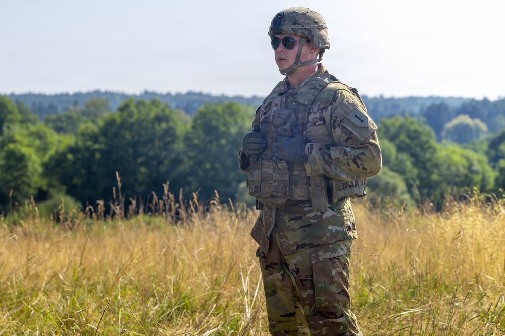 A U.S. Army soldier stands in an open field wearing full combat gear including OCP (Operational Camouflage Pattern) uniform, tactical vest, and combat helmet. The soldier appears to be observing or standing at attention in a rural outdoor environment with tall dry grass and a tree line in the background. The image appears to be taken during a training exercise or field operation.