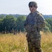 A U.S. Army soldier stands in an open field wearing full combat gear including OCP (Operational Camouflage Pattern) uniform, tactical vest, and combat helmet. The soldier appears to be observing or standing at attention in a rural outdoor environment with tall dry grass and a tree line in the background. The image appears to be taken during a training exercise or field operation.