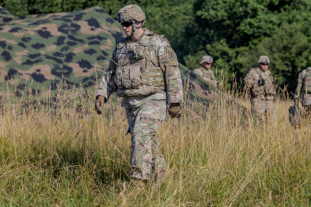 A U.S. Army soldier in full combat gear walks through a field of tall grass, with two additional soldiers visible in the background. The primary soldier wears Operational Camouflage Pattern (OCP) uniform, body armor, helmet with mounted device, and tactical gloves. A camouflage netting structure is visible in the background, suggesting a field training exercise or tactical operation.