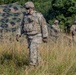 A U.S. Army soldier in full combat gear walks through a field of tall grass, with two additional soldiers visible in the background. The primary soldier wears Operational Camouflage Pattern (OCP) uniform, body armor, helmet with mounted device, and tactical gloves. A camouflage netting structure is visible in the background, suggesting a field training exercise or tactical operation.