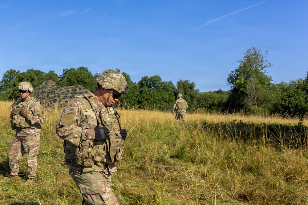 Three soldiers in MultiCam/OCP camouflage uniforms are operating in an open grassy field with tree lines in the background. A camouflaged military shelter or net structure is visible in the mid-left background. The scene appears to be a field training exercise or operational patrol in a temperate environment.