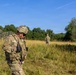 Three soldiers in MultiCam/OCP camouflage uniforms are operating in an open grassy field with tree lines in the background. A camouflaged military shelter or net structure is visible in the mid-left background. The scene appears to be a field training exercise or operational patrol in a temperate environment.