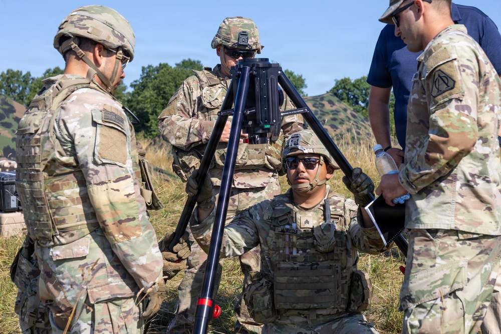 A group of U.S. Army soldiers in OCP (Operational Camouflage Pattern) uniforms are gathered around a tripod-mounted optical or targeting device in an outdoor field environment. One soldier is kneeling while operating or stabilizing the tripod-mounted equipment, while others observe or assist. The setting appears to be a training exercise based on the terrain and equipment configuration.