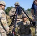 A group of U.S. Army soldiers in OCP (Operational Camouflage Pattern) uniforms are gathered around a tripod-mounted optical or targeting device in an outdoor field environment. One soldier is kneeling while operating or stabilizing the tripod-mounted equipment, while others observe or assist. The setting appears to be a training exercise based on the terrain and equipment configuration.