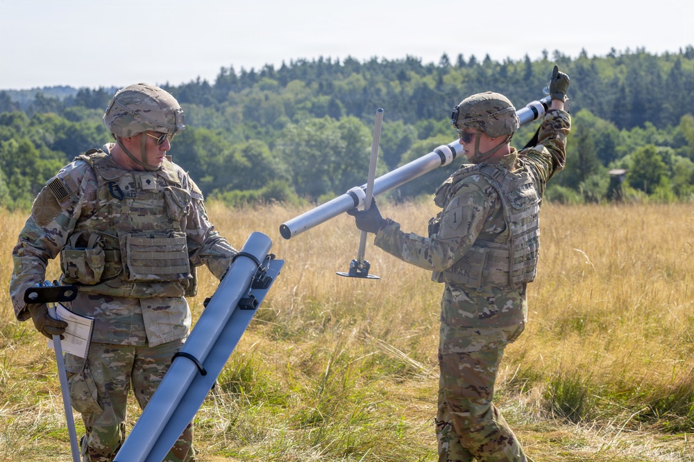 Two U.S. Army soldiers in MultiCam/OCP camouflage uniforms and combat gear are handling what appears to be a mortar system or launch tube assembly in an open field. They are working together to position or assemble the tubular equipment, with a treeline visible in the background. The setting appears to be a training area or exercise range during daylight hours.