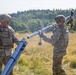 Two U.S. Army soldiers in MultiCam/OCP camouflage uniforms and combat gear are handling what appears to be a mortar system or launch tube assembly in an open field. They are working together to position or assemble the tubular equipment, with a treeline visible in the background. The setting appears to be a training area or exercise range during daylight hours.
