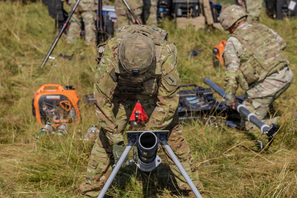 Military personnel in MultiCam/OCP camouflage uniforms are setting up equipment in a grassy field environment. The foreground shows a soldier leaning over what appears to be a mortar tube mounted on a bipod stand. Additional soldiers and equipment, including a portable generator, are visible in the background.