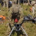 Military personnel in MultiCam/OCP camouflage uniforms are setting up equipment in a grassy field environment. The foreground shows a soldier leaning over what appears to be a mortar tube mounted on a bipod stand. Additional soldiers and equipment, including a portable generator, are visible in the background.