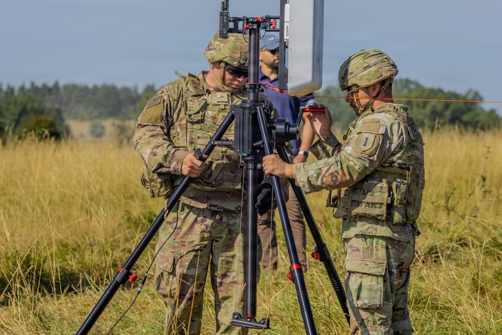 Two U.S. Army soldiers in OCP (Operational Camouflage Pattern) uniforms are setting up what appears to be a communications or sensor antenna mounted on a heavy-duty tripod in an open field. A civilian observer in a blue cap and brown pants stands in the background. The setting appears to be a field training exercise or equipment demonstration.