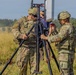 Two U.S. Army soldiers in OCP (Operational Camouflage Pattern) uniforms are setting up what appears to be a communications or sensor antenna mounted on a heavy-duty tripod in an open field. A civilian observer in a blue cap and brown pants stands in the background. The setting appears to be a field training exercise or equipment demonstration.