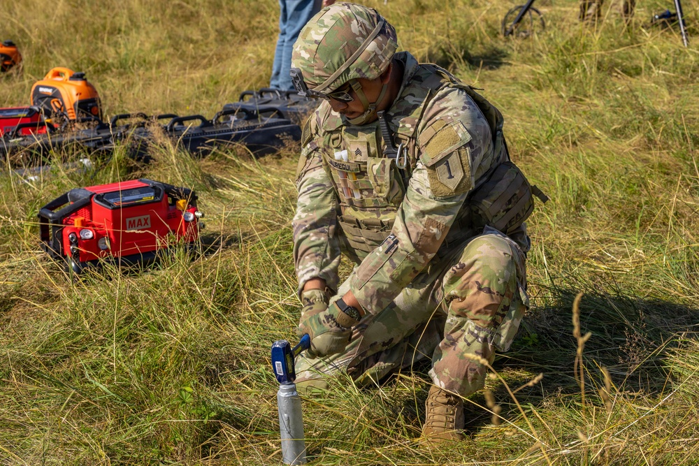A U.S. Army Sergeant in OCP (Operational Camouflage Pattern) uniform and body armor is kneeling in a grassy field, using a hammer to drive a stake or ground anchor into the earth. The soldier wears rank insignia consistent with Sergeant (three chevrons) and a unit patch on the left shoulder. Various pieces of equipment, including a red MAX-branded device and an orange generator, are visible in the background.