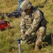 A U.S. Army Sergeant in OCP (Operational Camouflage Pattern) uniform and body armor is kneeling in a grassy field, using a hammer to drive a stake or ground anchor into the earth. The soldier wears rank insignia consistent with Sergeant (three chevrons) and a unit patch on the left shoulder. Various pieces of equipment, including a red MAX-branded device and an orange generator, are visible in the background.