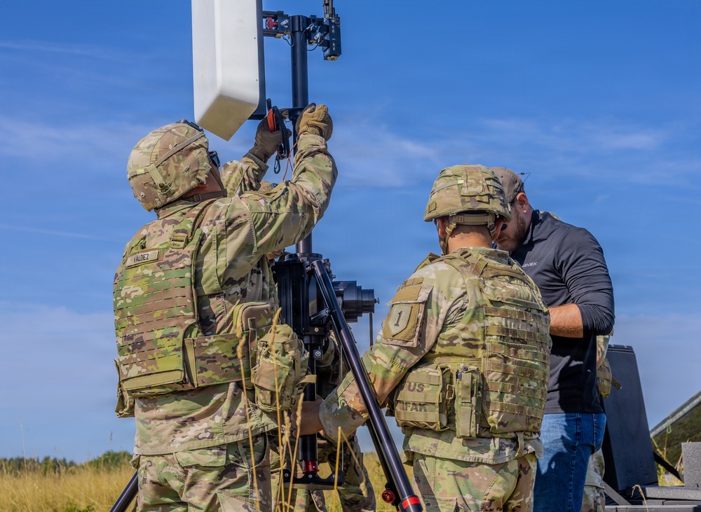 Two U.S. Army soldiers in OCP (Operational Camouflage Pattern) uniforms and body armor are assembling or adjusting a communications or sensor antenna mounted on a tripod in an open field. A civilian contractor in a dark polo shirt and jeans assists or observes the setup. The equipment appears to be a directional antenna or radar panel system mounted on a portable tripod stand.