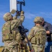 Two U.S. Army soldiers in OCP (Operational Camouflage Pattern) uniforms and body armor are assembling or adjusting a communications or sensor antenna mounted on a tripod in an open field. A civilian contractor in a dark polo shirt and jeans assists or observes the setup. The equipment appears to be a directional antenna or radar panel system mounted on a portable tripod stand.