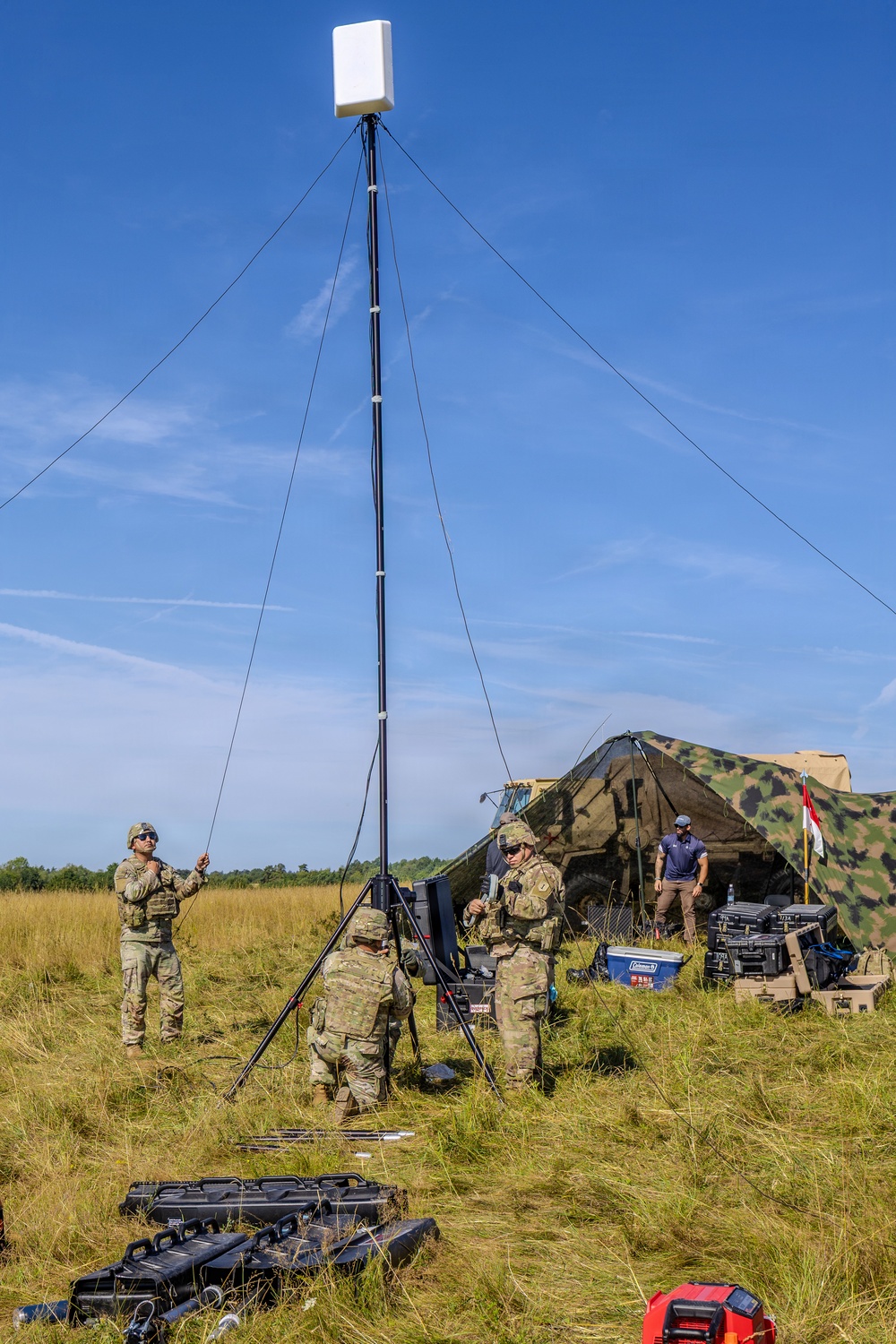 Military personnel in camouflage uniforms are setting up a portable communications or antenna mast in an open grassy field. The mast is stabilized with guy-wires and topped with a flat panel antenna or sensor unit. A camouflage-netted military tent structure and a civilian contractor are visible in the background alongside equipment cases.