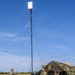 Military personnel in camouflage uniforms are setting up a portable communications or antenna mast in an open grassy field. The mast is stabilized with guy-wires and topped with a flat panel antenna or sensor unit. A camouflage-netted military tent structure and a civilian contractor are visible in the background alongside equipment cases.