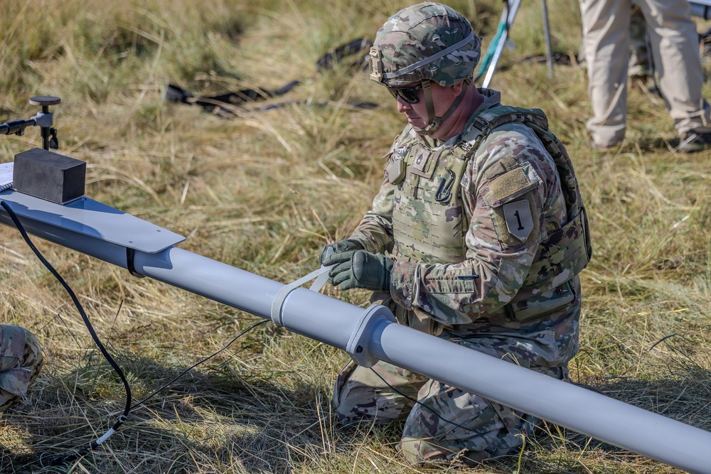 A U.S. Army soldier in full combat gear is kneeling on dry grassland, handling what appears to be a fixed-wing unmanned aerial vehicle (UAV) or drone component. The soldier is wearing Operational Camouflage Pattern (OCP) uniform with a unit patch showing the number '1' on the sleeve, tactical vest, helmet, and gloves. The setting appears to be an outdoor field exercise or UAV launch preparation.