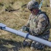 A U.S. Army soldier in full combat gear is kneeling on dry grassland, handling what appears to be a fixed-wing unmanned aerial vehicle (UAV) or drone component. The soldier is wearing Operational Camouflage Pattern (OCP) uniform with a unit patch showing the number '1' on the sleeve, tactical vest, helmet, and gloves. The setting appears to be an outdoor field exercise or UAV launch preparation.