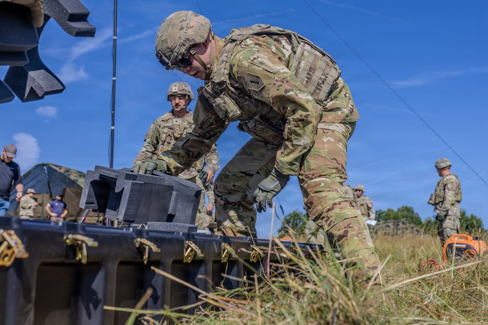 U.S. Army soldiers in Operational Camouflage Pattern (OCP) uniforms are working in an outdoor field environment, handling what appears to be military equipment cases or containers. The primary soldier in the foreground is leaning over a large black equipment case with yellow latches, appearing to open or inspect its contents. The setting suggests a field training exercise or equipment deployment operation.