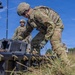 U.S. Army soldiers in Operational Camouflage Pattern (OCP) uniforms are working in an outdoor field environment, handling what appears to be military equipment cases or containers. The primary soldier in the foreground is leaning over a large black equipment case with yellow latches, appearing to open or inspect its contents. The setting suggests a field training exercise or equipment deployment operation.