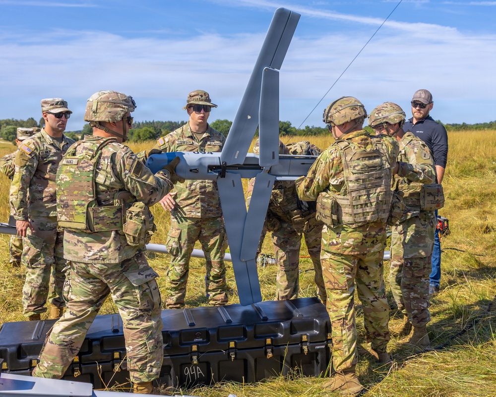 A group of U.S. Army soldiers in MultiCam/OCP camouflage uniforms are assembled around what appears to be a fixed-wing unmanned aerial vehicle (UAV/drone) in an open field. The personnel are engaged in what appears to be assembly, inspection, or pre-launch preparation of the aircraft. One civilian contractor in a dark polo shirt is also present among the military personnel.