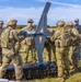 A group of U.S. Army soldiers in MultiCam/OCP camouflage uniforms are assembled around what appears to be a fixed-wing unmanned aerial vehicle (UAV/drone) in an open field. The personnel are engaged in what appears to be assembly, inspection, or pre-launch preparation of the aircraft. One civilian contractor in a dark polo shirt is also present among the military personnel.
