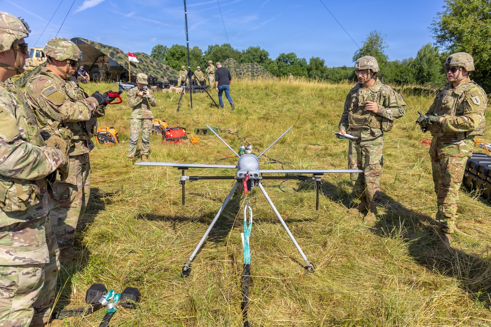 A group of U.S. Army soldiers in MultiCam/OCP camouflage uniforms are gathered around a large military unmanned aerial vehicle (UAV/drone) on a grassy field. The soldiers appear to be conducting a pre-flight inspection, briefing, or training exercise with the drone system. The setting appears to be a forward operating base or field exercise area, with camouflage netting, equipment, and communication antennas visible in the background.