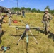 A group of U.S. Army soldiers in MultiCam/OCP camouflage uniforms are gathered around a large military unmanned aerial vehicle (UAV/drone) on a grassy field. The soldiers appear to be conducting a pre-flight inspection, briefing, or training exercise with the drone system. The setting appears to be a forward operating base or field exercise area, with camouflage netting, equipment, and communication antennas visible in the background.